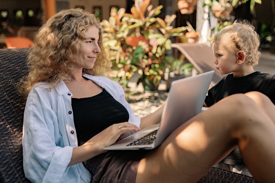 A woman working on a laptop outdoors with a child beside her on a sunlounger.