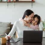 Happy young Asian woman working remotely from home with laptop and tablet while adorable little daughter hugging from behind