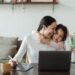Happy young Asian woman working remotely from home with laptop and tablet while adorable little daughter hugging from behind