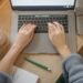 Top view of a woman typing on a laptop, surrounded by a coffee cup, notebook, and pen on a wooden table.