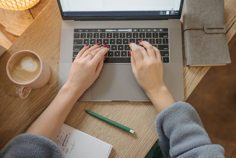 Top view of a woman typing on a laptop, surrounded by a coffee cup, notebook, and pen on a wooden table.