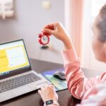 Woman holding a small clock at a desk with an open laptop screen displayed.