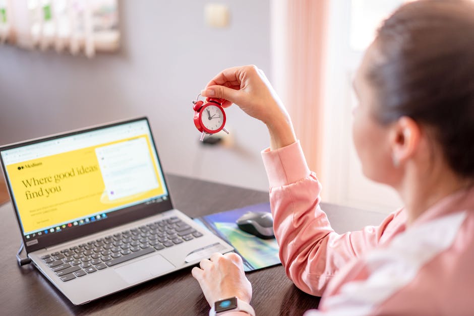 Woman holding a small clock at a desk with an open laptop screen displayed.
