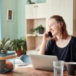 Smiling woman in a kitchen home office using a smartphone and laptop, surrounded by fruits.
