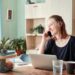 Smiling woman in a kitchen home office using a smartphone and laptop, surrounded by fruits.