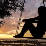 A serene sunset silhouette of a woman sitting on a swing by the seashore, evoking tranquility and reflection.