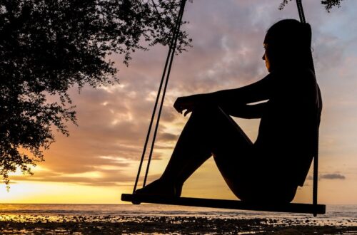 A serene sunset silhouette of a woman sitting on a swing by the seashore, evoking tranquility and reflection.