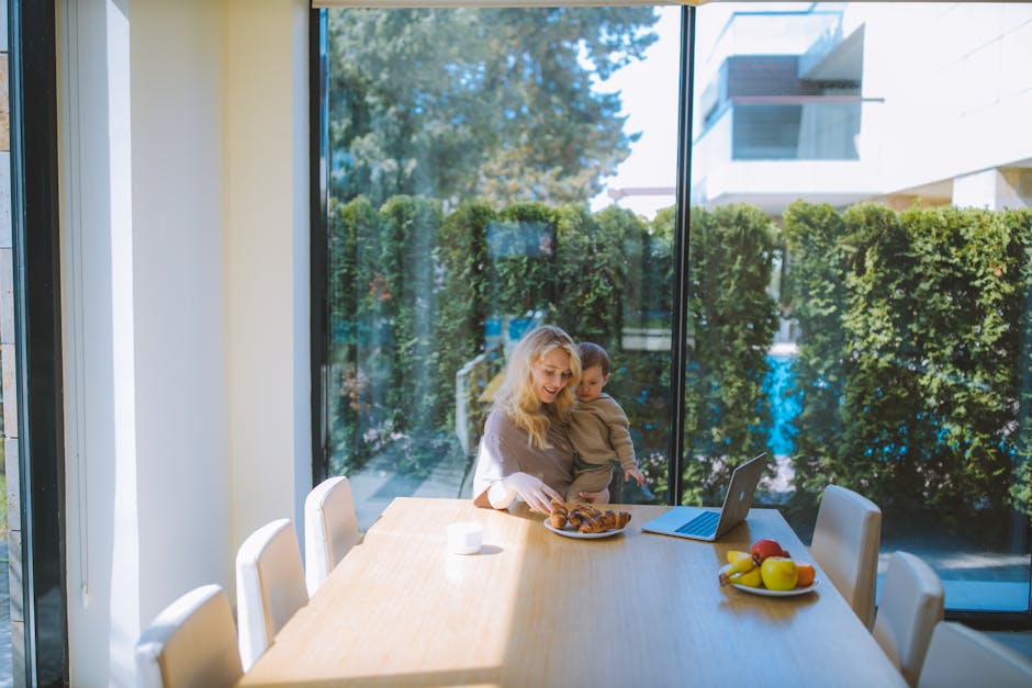 A mother multitasks with breakfast and remote work while holding her child at a dining table.